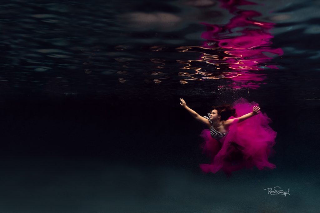 Underwater HS Senior in Pink Tulle and JCrew Navy Striped Suit Swimming in wide open deep blue pool with beautiful pink reflection